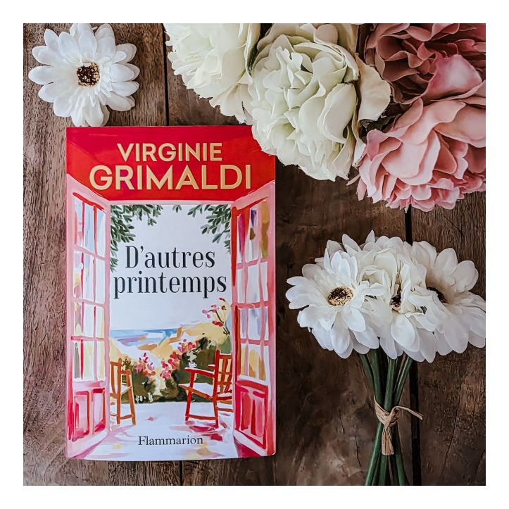 Roman D’autres printemps de Virginie Grimaldi posé sur une table en bois entouré de fleurs blanches et roses